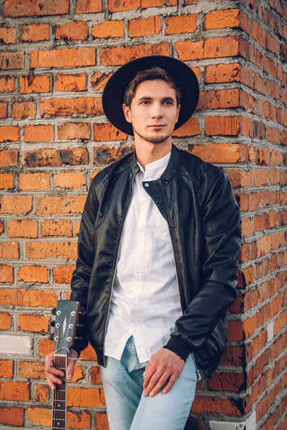 Young man with a guitar on the background of a brick wall. Handsome hippie style guitarist engrossed on music outdoors. Concept of freedom relaxation. - Photo, Image