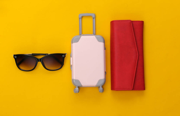 Voyage à plat. Mini valise de voyage en plastique, portefeuille rouge et lunettes de soleil sur fond jaune. Style minimal. Vue du dessus - Photo, image