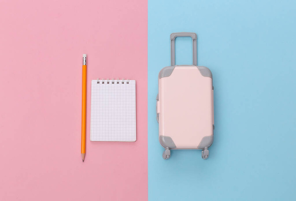 旅行計画。ミニプラスチック旅行スーツケースとノートブックピンクブルーパステルの背景。ミニマルなスタイル。トップビュー、フラットレイアウト - 写真・画像