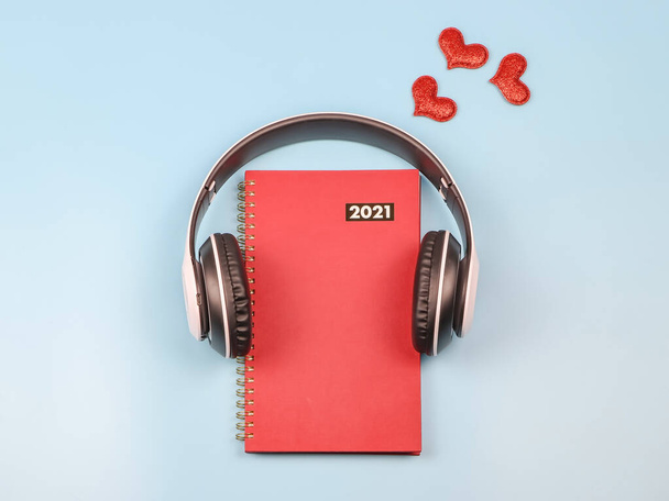 赤い輝きの心、コピースペースで飾られた青の背景にヘッドフォンで覆われた赤い2021日記やプランナーのトップビューまたはフラットレイアウト。2021年何を聴くべきか、オーディオブックの愛日記、バレンタインデー. - 写真・画像