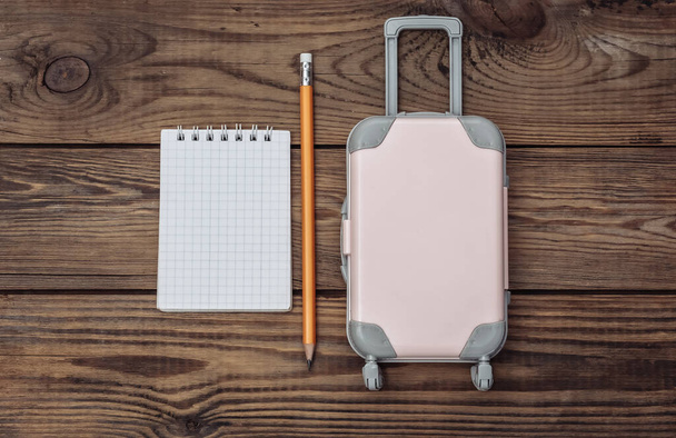 Płaskie wakacje lay i koncepcja strugania podróży. Mini plastikowa walizka podróżna i notatnik na drewnianym tle. Widok z góry - Zdjęcie, obraz