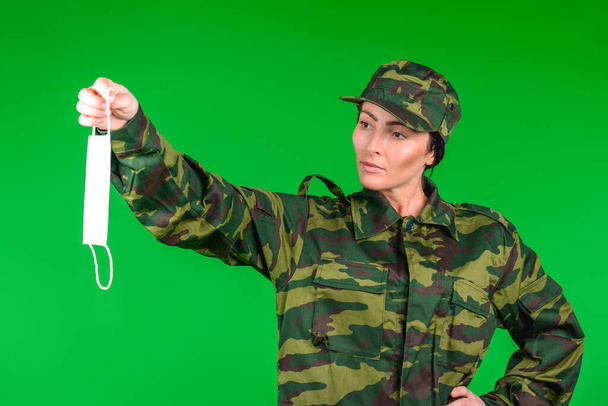 A serious woman in a military uniform holds out a medical mask on a green background. Gender neutrality in the covid 19 pandemic - Фото, зображення