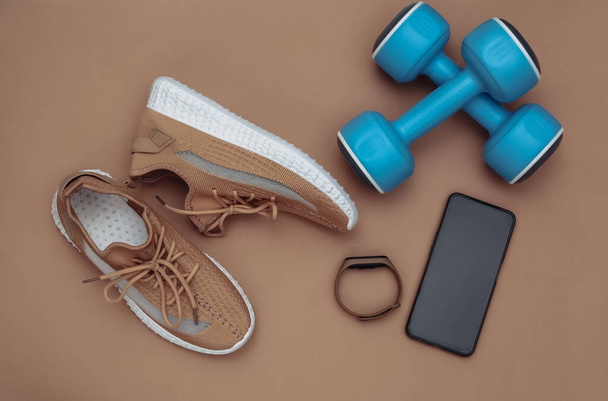 Hanteln, textile Sportschuhe, intelligentes Armband, Smartphone auf braunem Hintergrund. Fitnessflach lag. Ansicht von oben - Foto, Bild