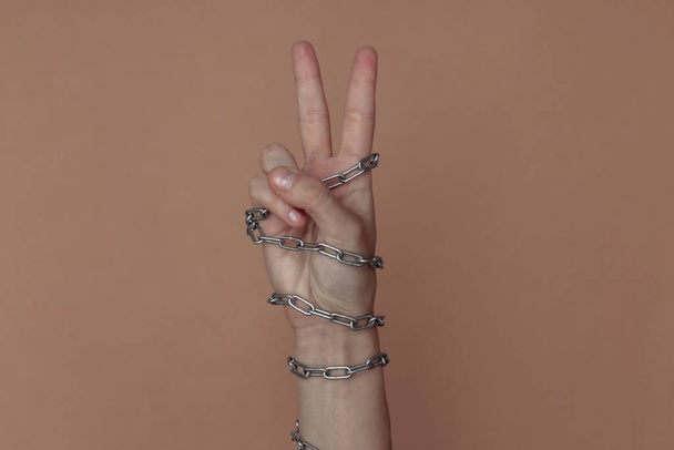 gesto de los dedos en V, mano envuelta en una cadena de metal sobre un fondo marrón. Supresión de la libertad o encarcelamiento - Foto, imagen