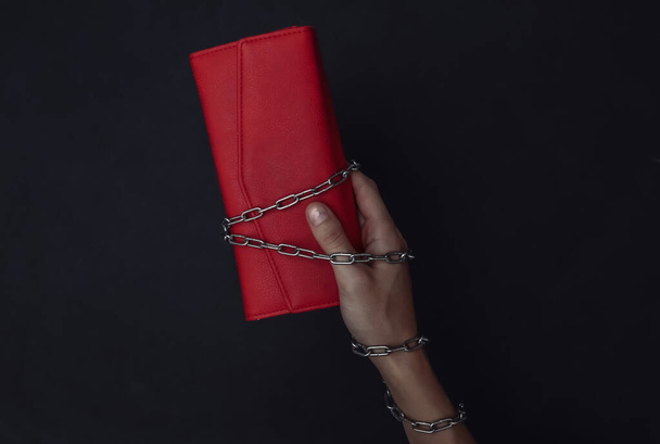 A hand wrapped in a steel chain holds a purse on black background. - Photo, Image