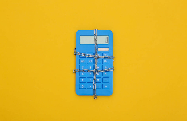 Голубой калькулятор, завернутый в стальную цепь на желтом фоне. Вид сверху - Фото, изображение