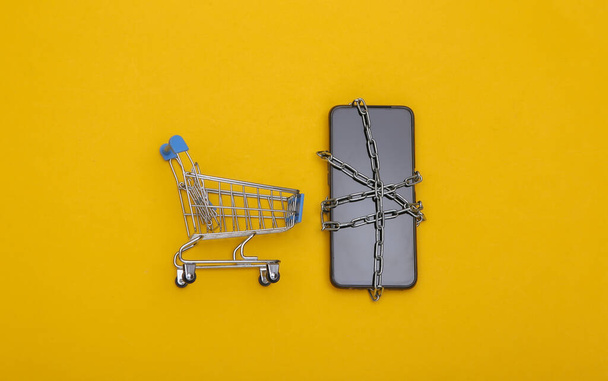 Smartphone wrapped in a steel chain an mini shoping trolley on yellow background. Top view - Foto, Imagen