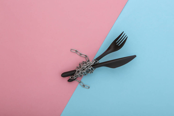 Tenedor de plástico y cuchillo envueltos en una cadena de acero sobre fondo pastel azul rosado. Concepto de dieta - Foto, Imagen