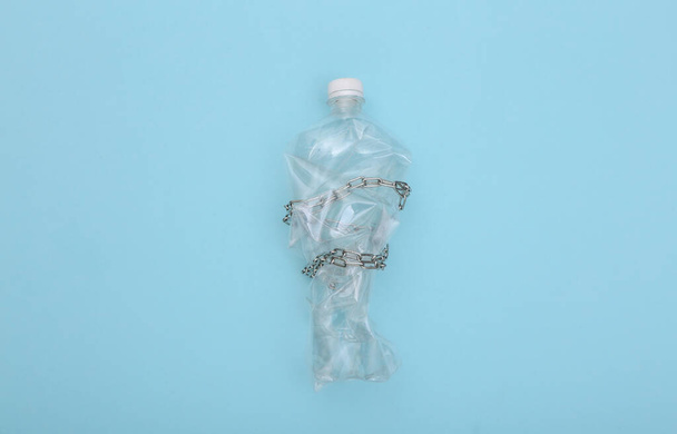 Garrafa de plástico amassada embrulhada em uma corrente de aço em um fundo azul. Conceito ecológico. Vista superior - Foto, Imagem