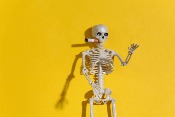 Σκελετός που καπνίζει με τσιγάρο στο στόμα σε ανοιχτό κίτρινο φόντο. Το κάπνισμα σκοτώνει - Φωτογραφία, εικόνα