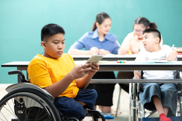 Ασιατικά ανάπηρα παιδιά (χαμένα πόδια) που χρησιμοποιούν tablet και κάθονται σε αναπηρικό καροτσάκι στην τάξη - Φωτογραφία, εικόνα
