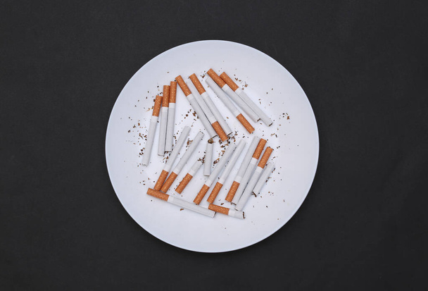 Many cigarettes in plate on black background. Top view - Zdjęcie, obraz