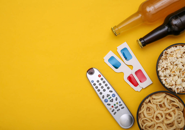 Filmzeit. Imbissschüsseln, Bierflaschen, Fernsehfernbedienung, 3D-Gläser auf gelbem Hintergrund. Ansicht von oben. Kopierraum - Foto, Bild