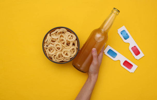 Movie time. Hand holds beer on yellow background with snacks and 3d glasses - Photo, Image