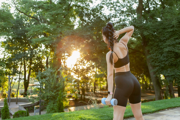 Привлекательная брюнетка-кавказка тренирует боковые мышцы живота с гантелями в руке в парке на рассвете. - Фото, изображение