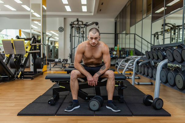 Tired muscular bodybuilder man sitting on a bench in the gym - 写真・画像