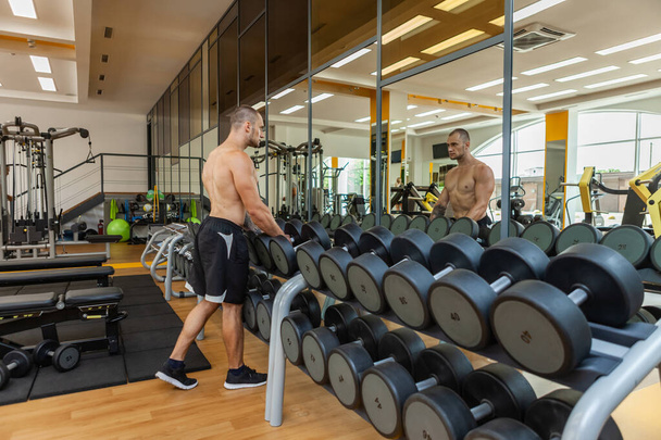 Muscular man with a naked torso removes dumbbells from a rack while looking in a mirror in a modern gym - Zdjęcie, obraz