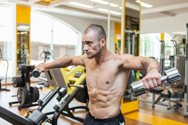 Beau musclé bodybuilder homme entraîne les muscles de l'épaule en soulevant haltères avec ses mains sur les côtés dans la salle de gym - Photo, image