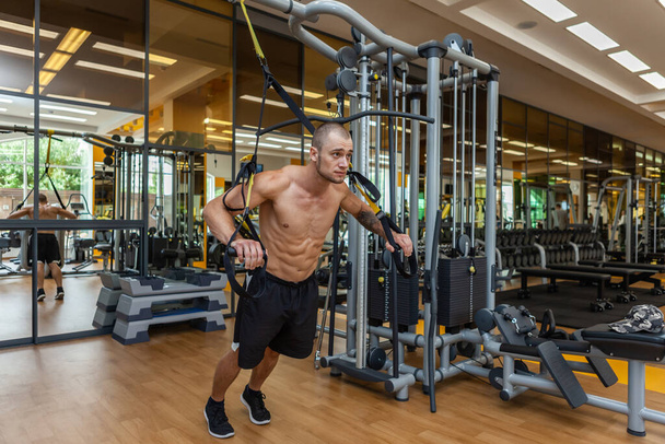 Svalnatý trup muž trénink s fitness trx systém v tělocvičně. Kulturista cvičí svaly se závěsnými popruhy - Fotografie, Obrázek