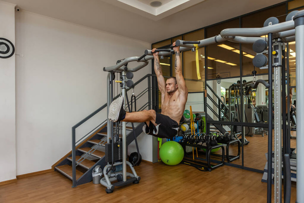 Young muscular fitness man training abdominal muscles while hanging on the horizontal bar in the gym - Photo, Image