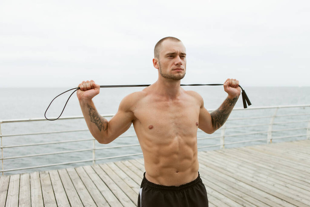 Geprägter muskulöser Mann mit nacktem Oberkörper und Springseil um den Hals am Strand - Foto, Bild