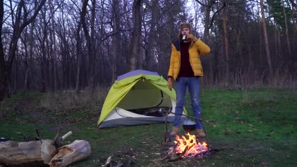 Um homem bebe chá perto de uma fogueira e uma tenda na floresta - Filmagem, Vídeo