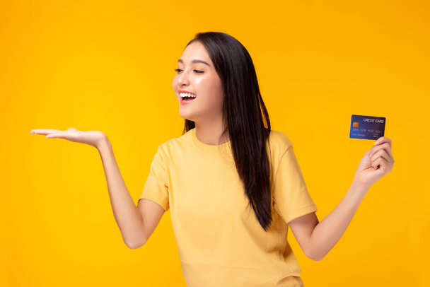 Happy young asian woman holding plastic credit card and looking at palm and copy space with satisfied Standing over yellow background Customer girl get happy of credit card service with smile on face - Zdjęcie, obraz
