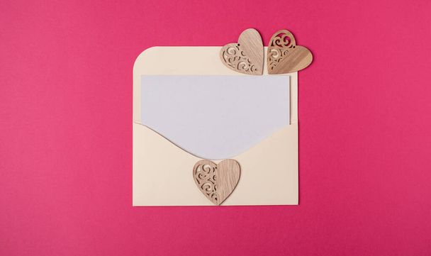 Envelope with a blank sheet of paper inside and wooden hearts on the hot pink background. Romantic love letter for the Valentine's day concept. Space for text. - Foto, Imagen