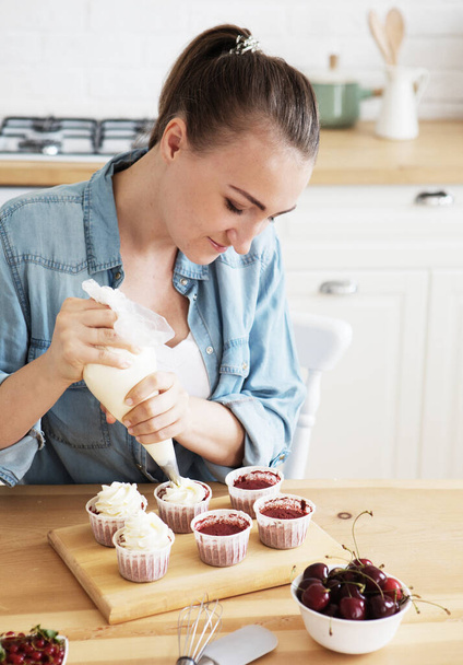 Lifestyle and people concept: A young woman decorates muffins with white cream in her kitchen. - Photo, Image