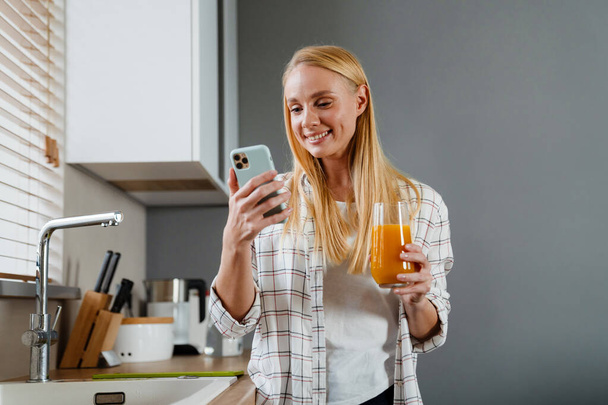 Blondynka szczęśliwa kobieta przy użyciu telefonu komórkowego i picia soku w kuchni domowej - Zdjęcie, obraz