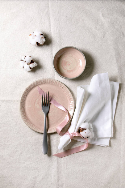 Set of empty pink ceramic plates with fork and textile napkin, decorated by cotton flowers and silk ribbon over white linen tablecloth. Table setting. Flat lay, space. - Φωτογραφία, εικόνα