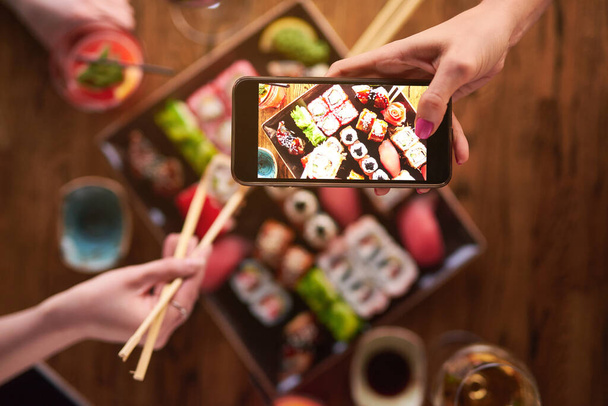Arriba de la vista. Dos chicas están comiendo sushi y tomar fotos en el teléfono inteligente. Conjunto de varios tipos de rollos y sushi con bebidas - Foto, imagen
