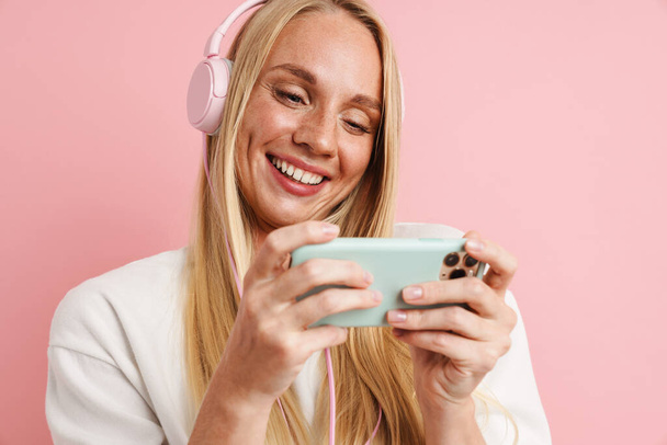 Cheerful beautiful girl in headphones playing video game on cellphone isolated over pink background - Foto, Imagen