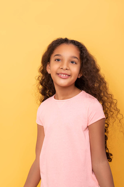 Waist up portrait of a dark skinned young girl with long curly hair wearing pink shirt on a yellow background - Фото, изображение