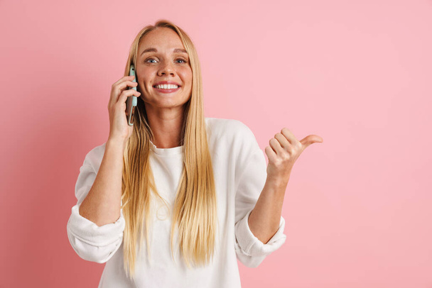 Joyful beautiful girl pointing finger aside and talking on cellphone isolated over pink background - Foto, Bild