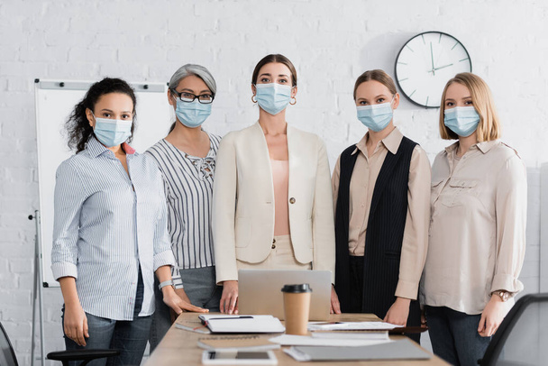 мультикультурные предпринимательницы в медицинских масках рядом с рабочим местом в конференц-зале - Фото, изображение