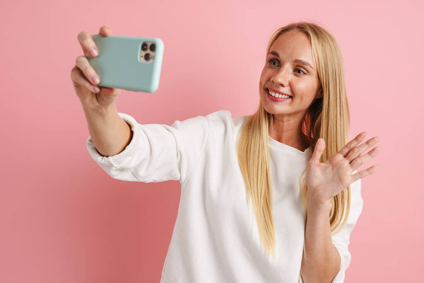 Joyful mooi meisje zwaaien hand terwijl het nemen van selfie op mobiele telefoon geïsoleerd over roze achtergrond - Foto, afbeelding