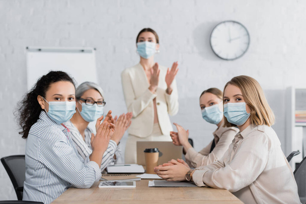 interracial businesswomen in medical masks applauding with team leader on blurred background - Fotografie, Obrázek