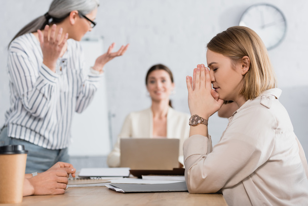 worried businesswoman with closed eyes and praying hands sitting at desk with interracial colleagues on blurred background  - Photo, Image