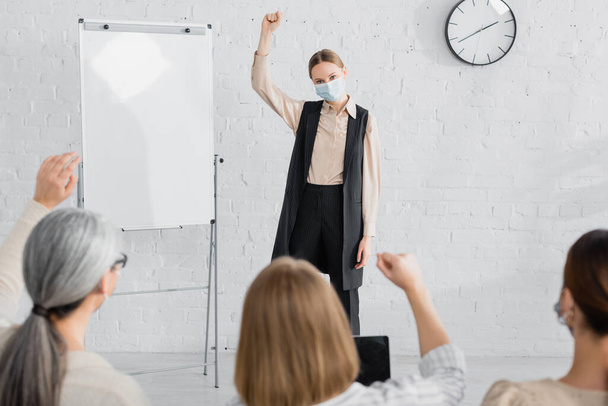 speaker in medical mask raising hand while standing near flipchart and looking at businesswomen on blurred foreground - Fotografie, Obrázek