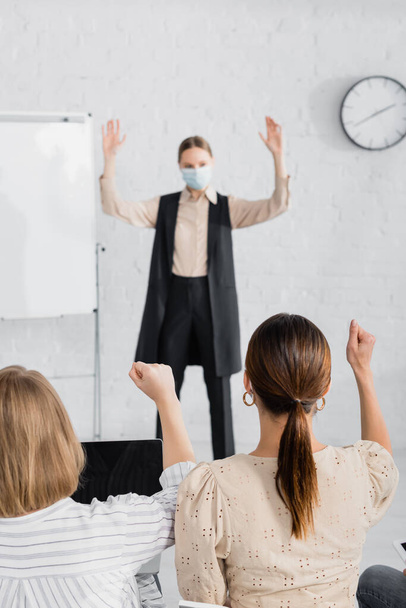 back view of businesswomen raising hands near speaker in medical mask standing near flipchart on blurred background  - 写真・画像