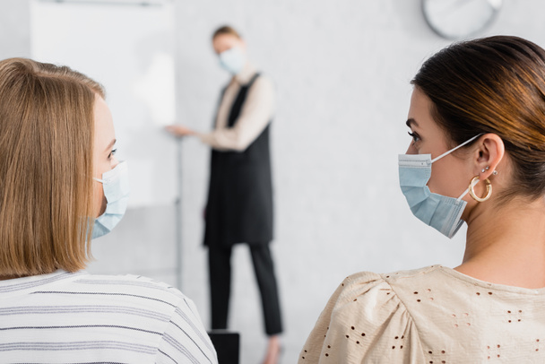 young businesswomen in medical masks looking at each other during seminar - Фото, изображение