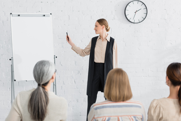 cheerful speaker looking at blank flipchart near woman during seminar on blurred foreground  - Foto, Imagem