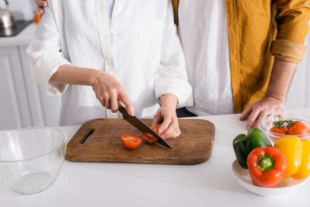 Rajatut näkymä vanhempi mies halaus vaimo leikkaus tomaatti lähellä vihanneksia ja kulhoon hämärtynyt etualalla  - Valokuva, kuva
