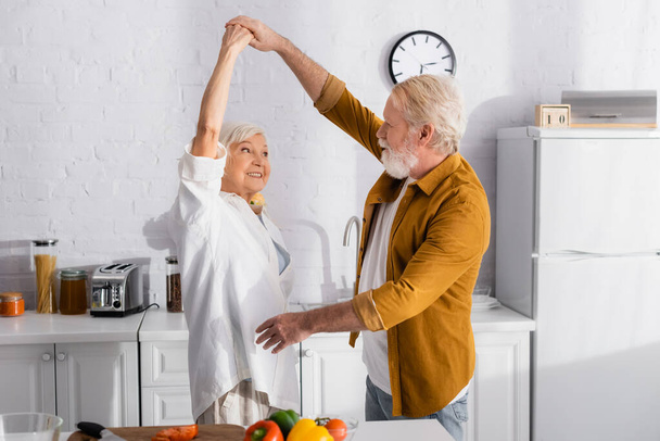 キッチンテーブルの上で新鮮な野菜の近くで踊る陽気なシニアカップル  - 写真・画像