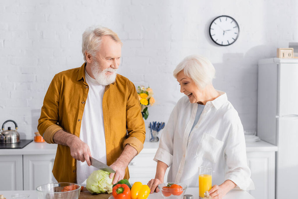 Man cutting cabbage near fresh vegetables and smiling wife with orange juice in kitchen  - Foto, Bild