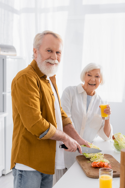 Starszy mężczyzna patrzy w kamerę podczas krojenia warzyw w pobliżu uśmiechniętej żony szklanką soku pomarańczowego na rozmazanym tle w kuchni  - Zdjęcie, obraz