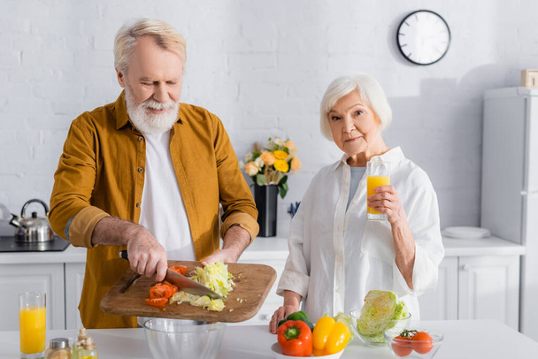 Senior woman holding glass of orange juice near husband pouring vegetables in bowl in kitchen  - Fotoğraf, Görsel