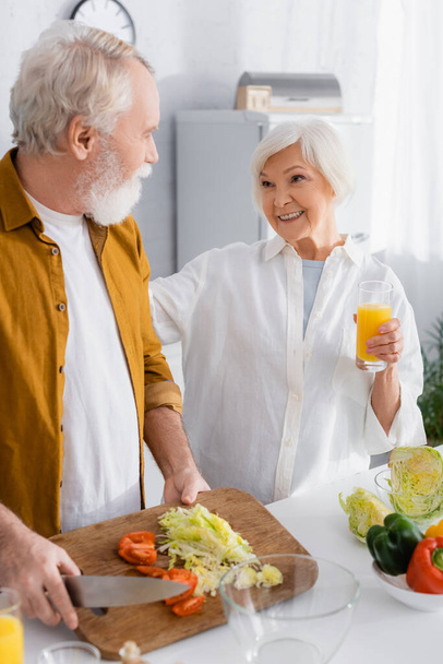 glimlachende senior vrouw met sinaasappelsap op zoek naar echtgenoot snijden groenten in de keuken  - Foto, afbeelding