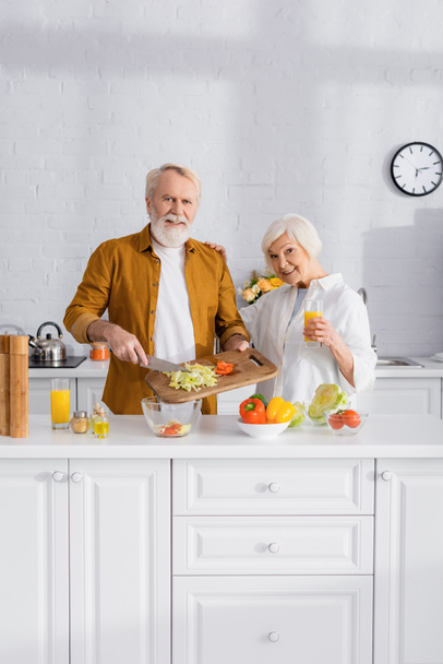 Vidám idős pár narancslével főzés a konyhaasztalon  - Fotó, kép